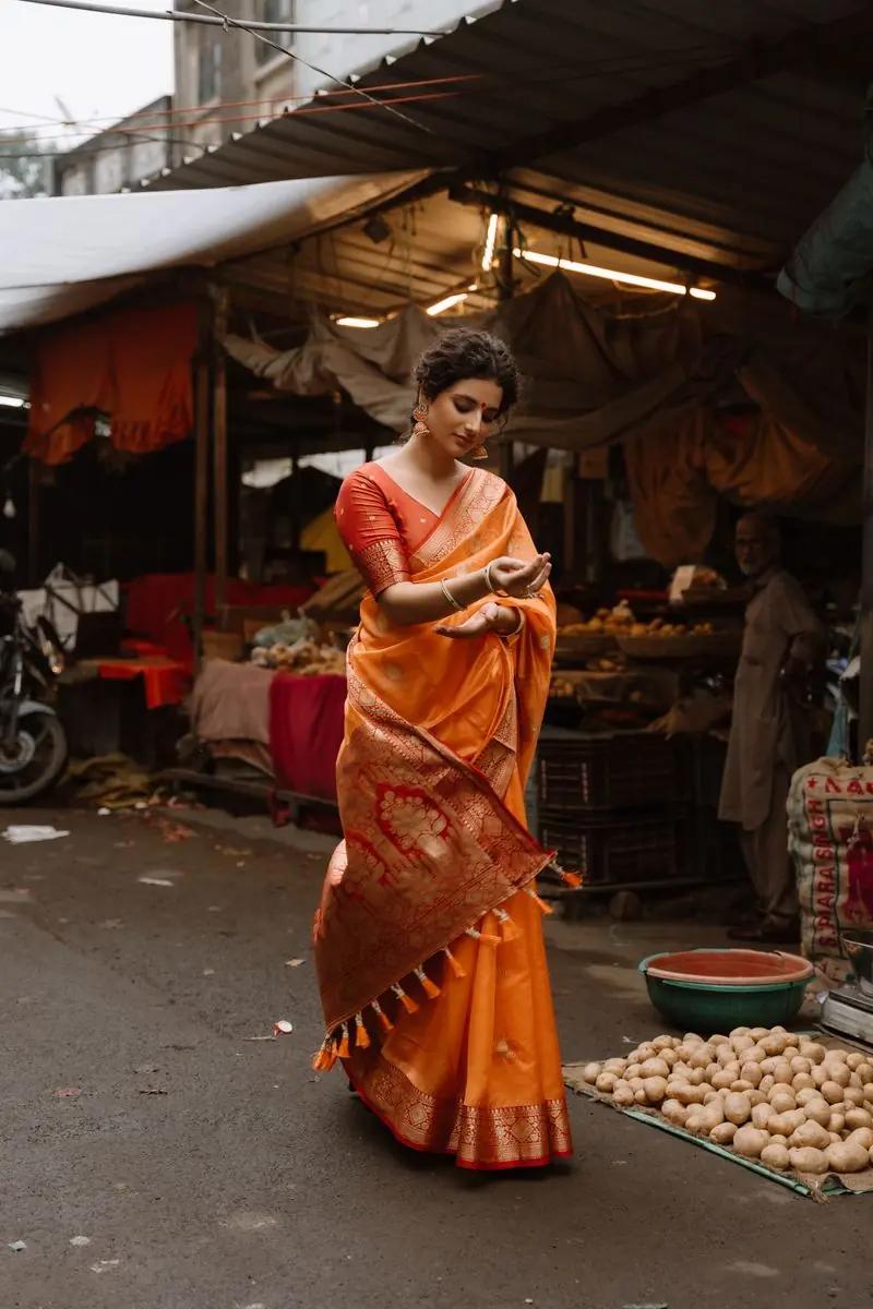 A woman in an orange Banarasi saree drapes the pallu in a busy market lane